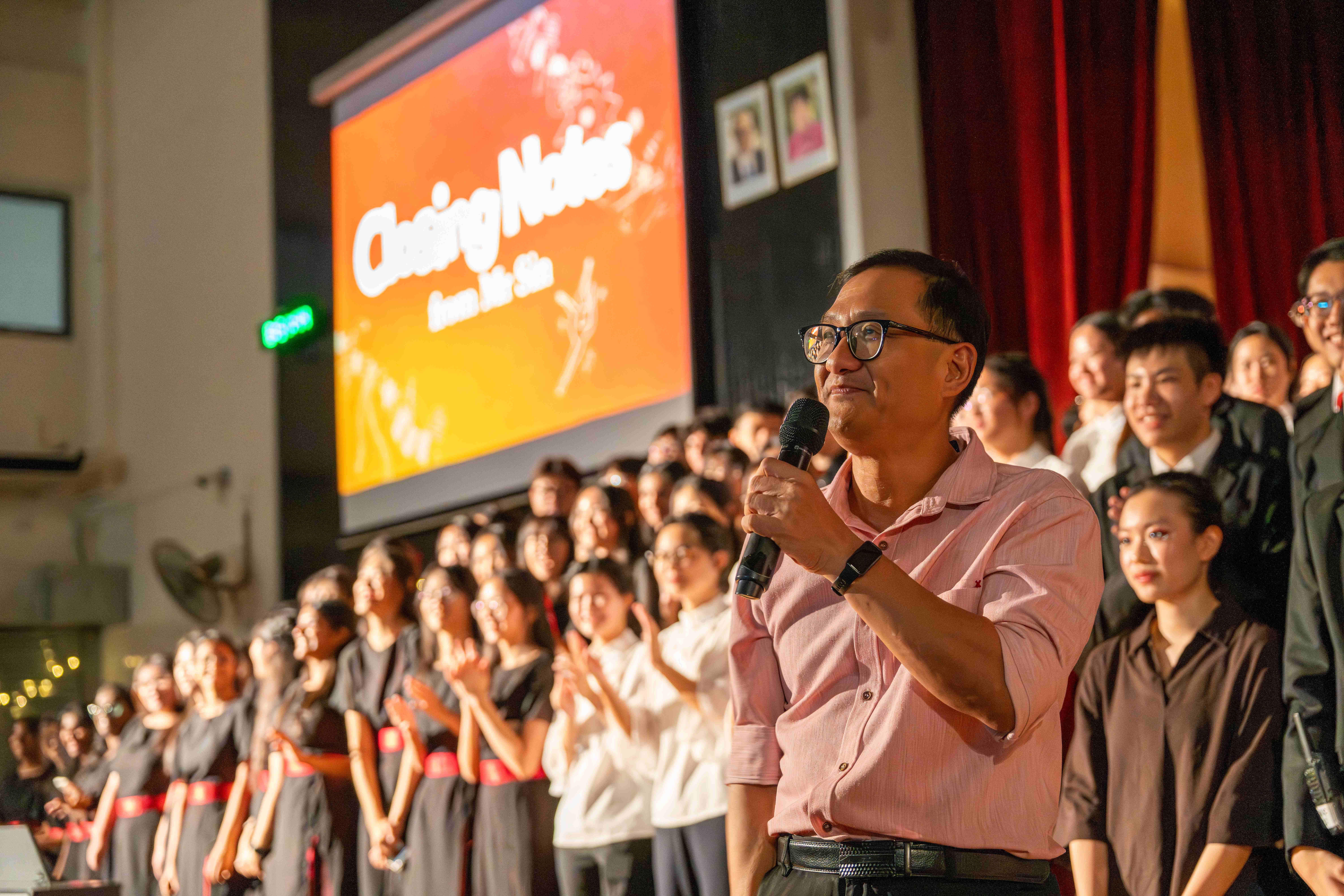 A man in a pink shirt speaks into a microphone on stage during "Closing Notes", with rows of students standing and applauding behind him.